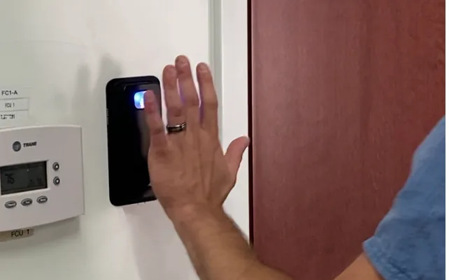 Hand held over a wall-mounted palm vein reader beside a door